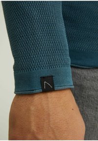 Close-up of a wrist wearing a textured blue sleeve with a small black fabric tag stitched near the cuff.