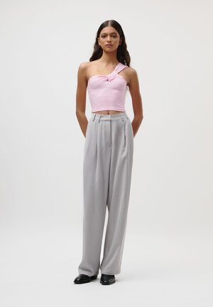 Vrouw met lang donker haar draagt een roze crop top met één schouder, lichtgrijze, hooggetailleerde broek met wijde pijpen en zwarte schoenen, staand op een witte achtergrond.