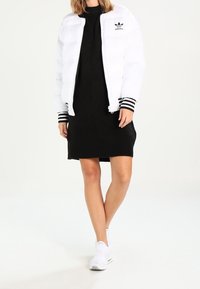 Donna che indossa un giubbotto imbottito bianco Adidas con polsini a strisce nere sopra un abito nero e sneakers bianche senza lacci, in piedi con le gambe incrociate.