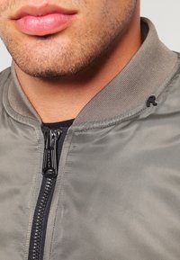 Šedá bombardérka se zipovým zapínáním, žebrovaným límcem a černým zipem. Obsahuje malé černé logo poblíž límce na pravé straně.