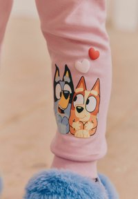 Różowe bawełniane legginsy z nadrukiem dwóch kreskówkowych psów po bokach, z trzema małymi kolorowymi serduszkami nad nimi. Miękkie niebieskie futrzane obuwie.