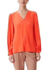 Blouse orange vif avec un col en V, manches longues avec des poignets froncés et une texture lisse et légère. Portée sur un pantalon à carreaux.