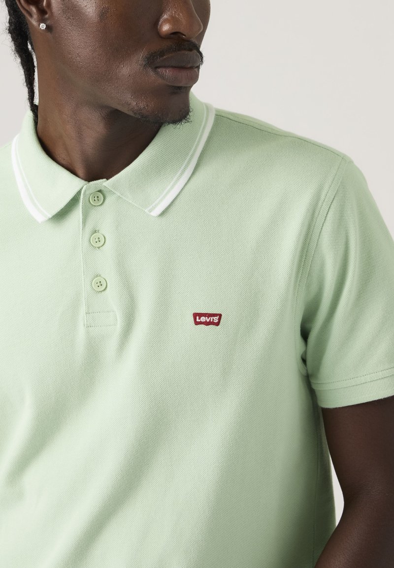Mann trägt ein hellgrünes Levi's Poloshirt mit weißen Kragenstreifen, drei Knöpfen geschlossen und rotem Levi's Logo auf der Brust.