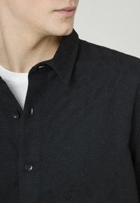 Homme portant une chemise noire texturée à boutons sur un t-shirt blanc, regardant vers la gauche, vue en gros plan du cou et du haut de la poitrine.