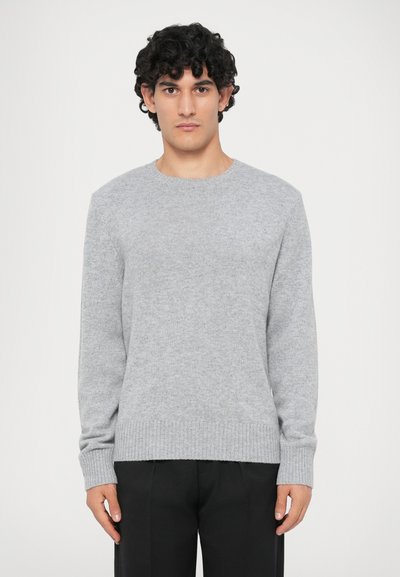 Homme avec des cheveux bouclés portant un pull en laine gris clair et un pantalon noir, se tenant devant un fond blanc uni.