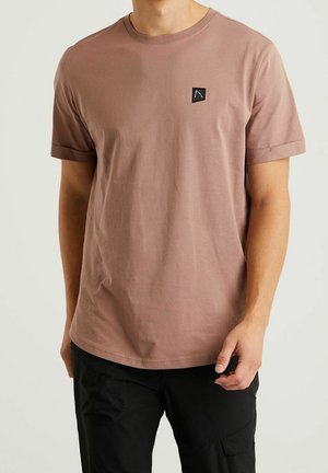 Koszulka krótki rękaw, okrągły dekolt w stonowanym różowym kolorze z bawełny. Posiada małą czarną naszywkę z logo po lewej stronie klatki piersiowej. Prosty dół i podwinięte rękawy.