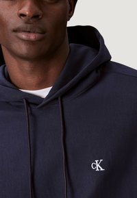 Marinblå hoodie i mjukt tyg, med en vit broderad "ck"-logotyp på bröstet och en justerbar huva. Slät textur.