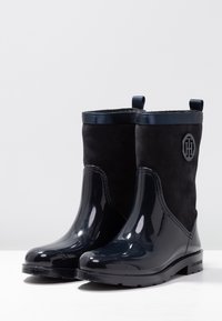 Mustat kumisaappaat kiiltävällä pinnalla ja nupukkinahkaisella päällisellä. Niissä on kaartuva muotoilu, lenkkikahvat ja logo sivussa.