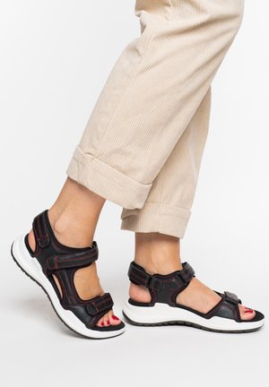 Person wearing beige rolled-up pants and black sporty sandals with white soles and red stitching, toes painted red, stepping forward.