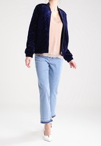 Giacca bomber trapuntata blu navy con polsini a coste, indossata sopra un top rosa chiaro con pizzi, abbinata a jeans azzurri e tacchi bianchi.