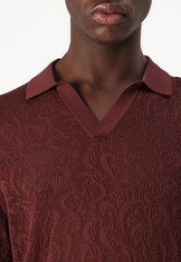Polo shirt bordeaux foncé avec un motif en maille texturée, un design encolure en V et un col côtelé. Tissu lisse avec un éclat subtil.
