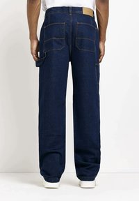 Mörkblå denimbyxor med avslappnad passform, med flera fickor, stickade detaljer och en enkel läderpatch vid midjan.