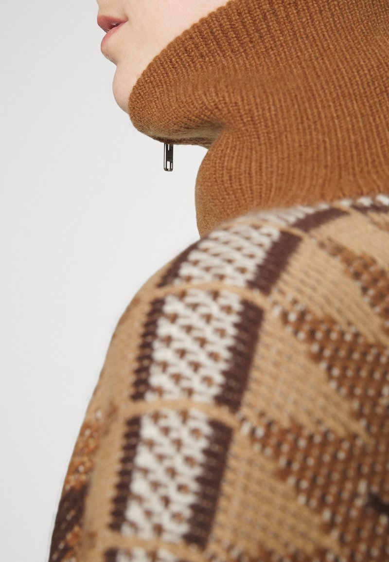 Brauner gestrickter Pullover mit hohem Kragen, der geometrische Muster in Creme und Dunkelbraun zeigt, strukturierte Oberfläche, sichtbares Reißverschlussdetail.