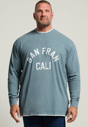 Homme aux cheveux courts et à la barbe portant une chemise bleue à manches longues avec le texte "SAN FRAN CALI" sur un fond clair uni.