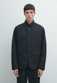 Sort jakke med høj krave, knaplukning og to forlommer. Glat stof med afslappet pasform, skræddersyet silhuet og ingen synlige mønstre.