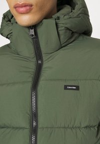 Doudoune verte avec col montant et capuche. Dotée d'une fermeture éclair noire sur toute la longueur et d'une petite étiquette logo Calvin Klein sur la poitrine.