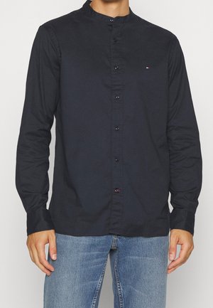 Mand iført navyblå skjorte med mandarin-krave med knapper og lange ærmer samt lyseblå jeans mod ensfarvet baggrund.