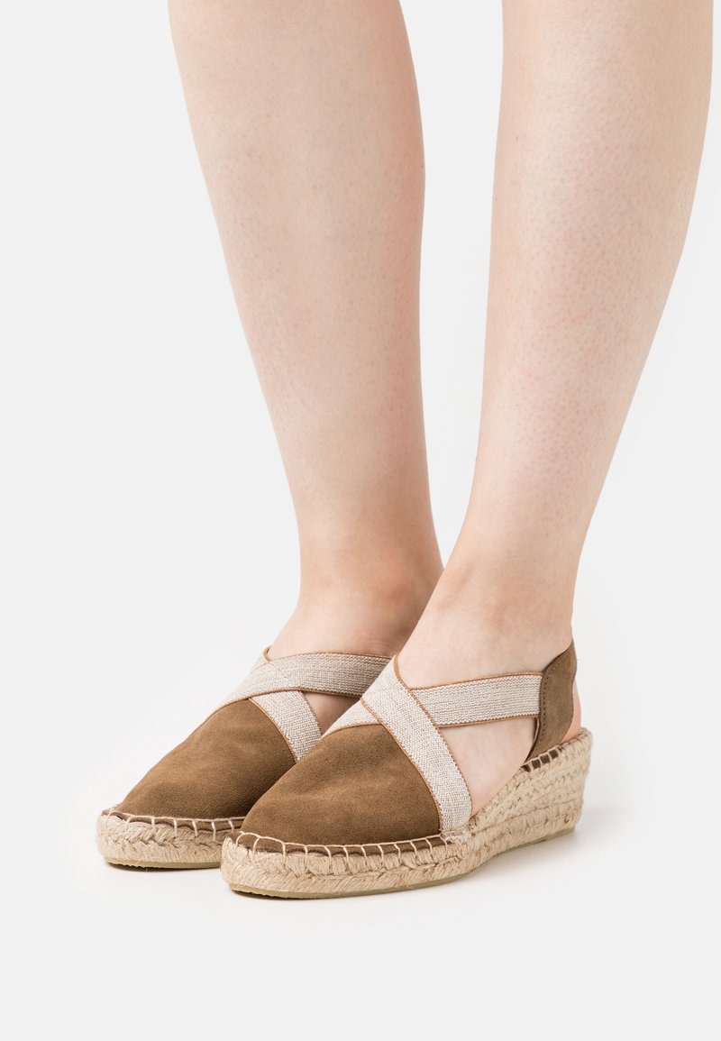 Espadrilles compensées en daim marron avec des brides élastiques croisées beige portées pieds nus sur fond blanc.