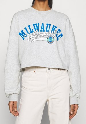 Osoba nosząca jasnoszary krótki sweter z napisem "Milwaukee Wisconsin" i logo softbolu, w połączeniu z wysokim stanem w kolorze ecru.