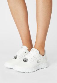 Baskets de sport blanches avec une texture lisse, un design à lacets et une semelle flexible. Présente un logo et une étiquette antidérapante sur le côté.