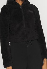 Svart kort zip-up hoodie i fluffigt material med dragsko i nederkant och en liten broderad logotyp på bröstet.