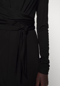 Schwarzes Langarmkleid mit drapiertem Design und gebundenem Taillendetail. Glatte Stofftextur und figurbetonte Ärmel.