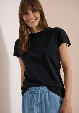 Femme souriante aux longs cheveux bruns portant un haut noir à manches courtes et un pantalon bleu clair à cordon, sur un fond uni.