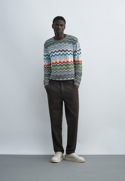 Άνδρας που στέκεται μπροστά σε λευκό τοίχο, φοράει πολύχρωμο πουλόβερ με κυματιστό σχέδιο, σκούρο καφέ παντελόνι και λευκά αθλητικά παπούτσια με τα χέρια στις τσέπες.