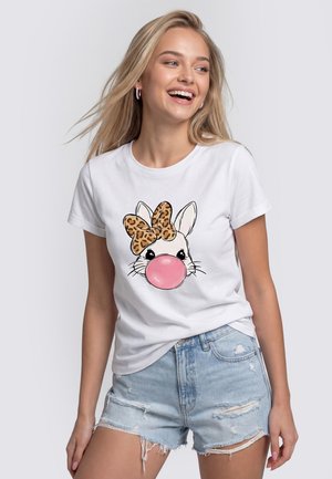 Junge Frau trägt weißes T-Shirt mit Hasengesicht, bläst rosa Kaugummiblase und trägt eine Leopardenmuster-Schleife, kombiniert mit hellen, zerrissenen Jeansshorts.