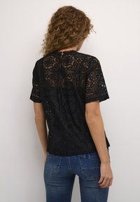 Black lace top featuring intricate floral patterns, short sleeves, and a button closure at the back. Worn with blue jeans.