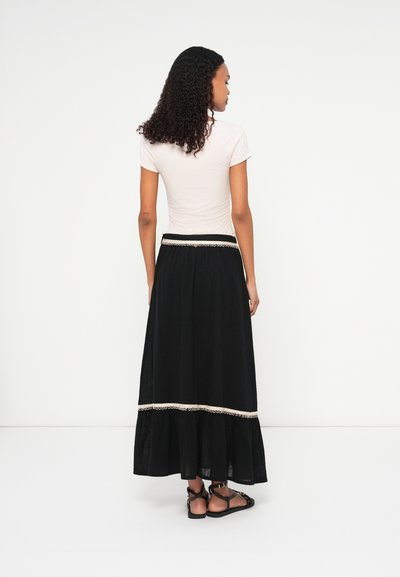 Črna maksi krila s prosojnim dizajnom, ki ima čipkast rob na dnu in prilegajoč pas. V kombinaciji z svetlo obarvano majico in sandali.