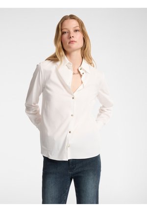 Camisa blanca de botones con cuello, que presenta una tapeta frontal, mangas largas y un ligero brillo; combinada con jeans de mezclilla oscura.