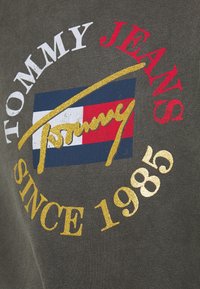 Šedá bavlněná tkanina s potiskem značky v bílé, červené a zlaté. Obsahuje logo s designem vlajky a text "SINCE 1985" pod ním.