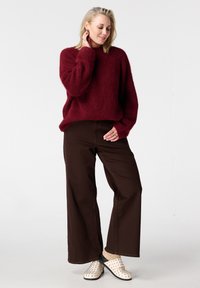 Pull en laine bordeaux avec un effet duveteux, associé à un pantalon large marron. Chaussures blanches à enfiler avec des accents cloutés. Design minimaliste et texture douce.