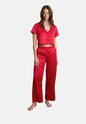Femme debout pieds nus portant un crop top rouge en satin à manches courtes boutonné et un pantalon large assorti à pois blancs sur fond blanc.
