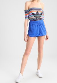 Multicolored striped off-shoulder top with an Adidas logo, paired with blue drawstring shorts. Model wearing white sneakers.