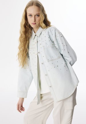 Giacca di jeans azzurro chiaro con borchie argentate, una tasca frontale e chiusura con bottoni. Presenta una vestibilità comoda e una texture morbida.