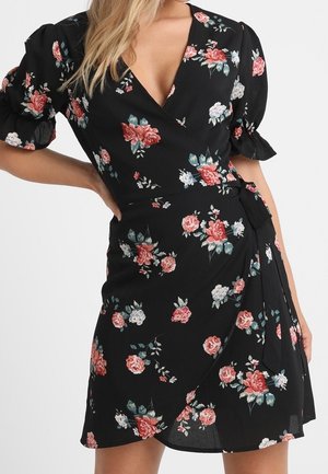 Zwarte wikkeljurk met pofmouwen en roze, wit en groen bloemenpatroon, gedragen met hand op de heup.