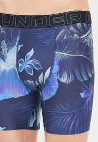 Boxeri scurți cu imprimeu floral albastru, având un tiv elastic negru cu textul "UNDER ARMOUR". Materialul are un finisaj neted și texturat.