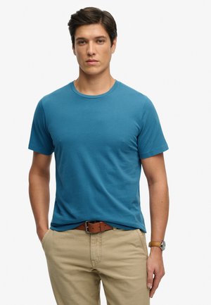 Joven con cabello oscuro que lleva una camiseta azul de manga corta, pantalones beige, cinturón marrón y reloj de pulsera, de pie con una mano en el bolsillo.