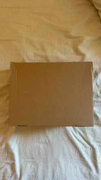 Brown rectangular cardboard box with a smooth texture, slightly creased edges, resting on a light-colored fabric surface.