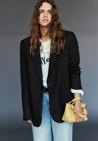 Mlada oseba z dolgimi valovitimi lasmi nosi prevelik črn blazer, belo majico s potiskom, svetlo modre kavbojke ter drži majhno rumeno torbico na sivi podlagi.