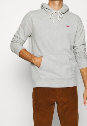 Homme portant un sweat à capuche gris clair avec poche avant et un pantalon en velours côtelé marron, la main placée dans la poche du sweat, fond blanc.