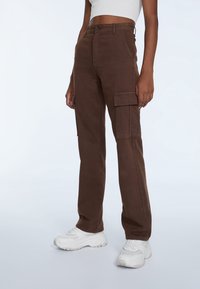 Heren bruine cargobroek met een hoge taille, grote zijzakken en een rechte pijpdesign. Materiaal lijkt stevig met een gladde textuur.