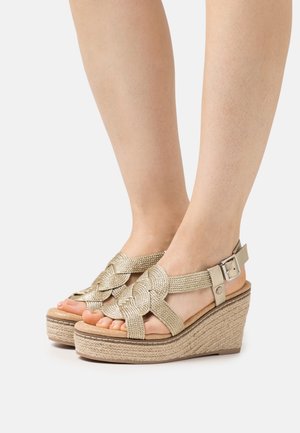 Sandały na koturnie z plecionego metalicznego materiału, beżowe paski, otwarty czubek i strukturalna, pleciona podeszwa. Regulowany pasek na kostkę z klamrą.