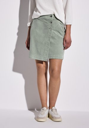 Jupe en denim vert clair avec une fermeture à boutons diagonale à l'avant, ajustement sur mesure et texture douce. Portée avec des baskets blanches pour le contraste.