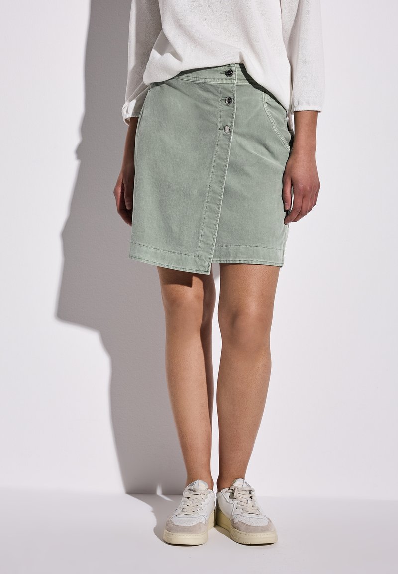 Helles grünes Denim-Rock mit diagonaler Knopfleiste vorne, taillierter Passform und zarter Textur. Kombiniert mit weißen Sneakers für einen Kontrast.