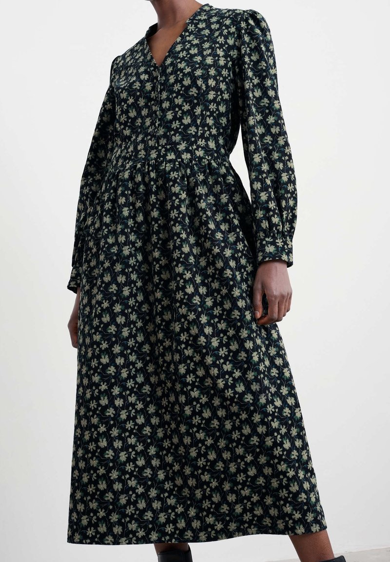 Femme portant une robe midi à manches longues, imprimé floral foncé, col en V et taille froncée sur un fond clair uni.