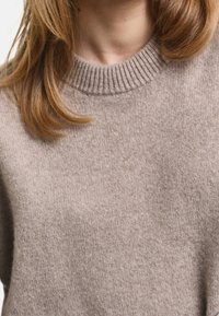 Beżowy sweter wykonany na drutach, z prążkowanym okrągłym dekoltem i miękką, teksturowaną powierzchnią. Materiał wydaje się ciepły i uniwersalny do warstwowego ubierania.