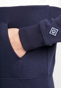 Sudadera azul marino con textura acanalada, que cuenta con un bolsillo frontal y un pequeño parche con el logo en la manga. Tejido suave y confortable.